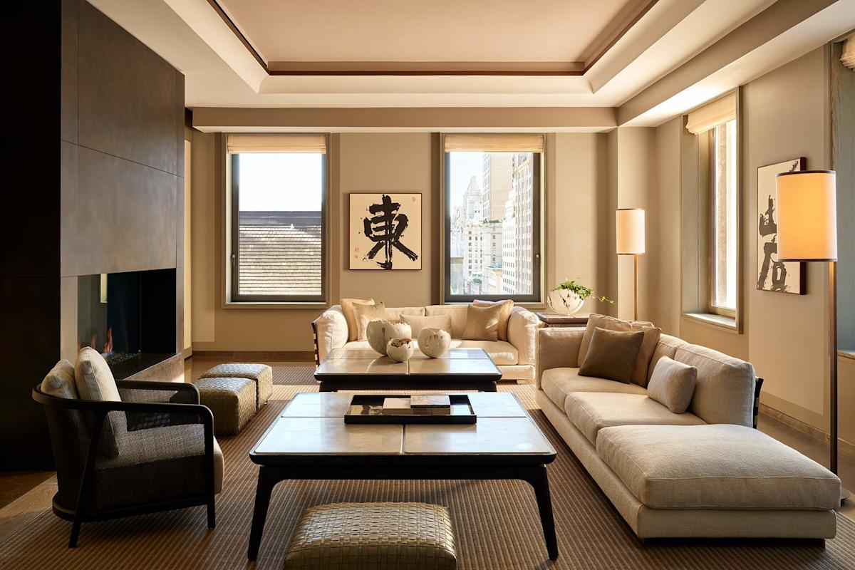 A modern living room with beige sofas, a black armchair, a fireplace, large windows, a low coffee table, neutral decor, and Asian-inspired wall art. Natural light fills the elegant, spacious space.