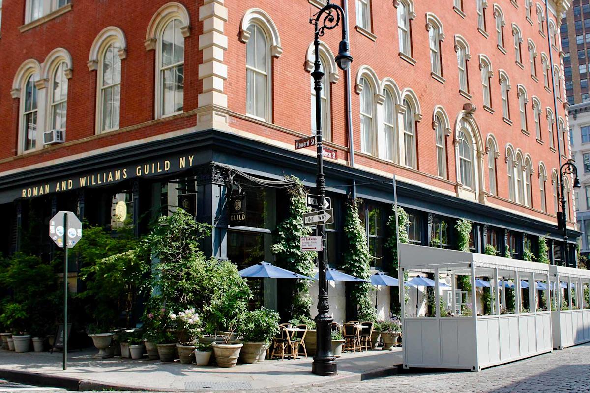 Street view of a red-brick corner building with arched windows, a black storefront labeled "Roman and Williams Guild NY," outdoor dining area with blue umbrellas, greenery, and street signs on a sunny day.