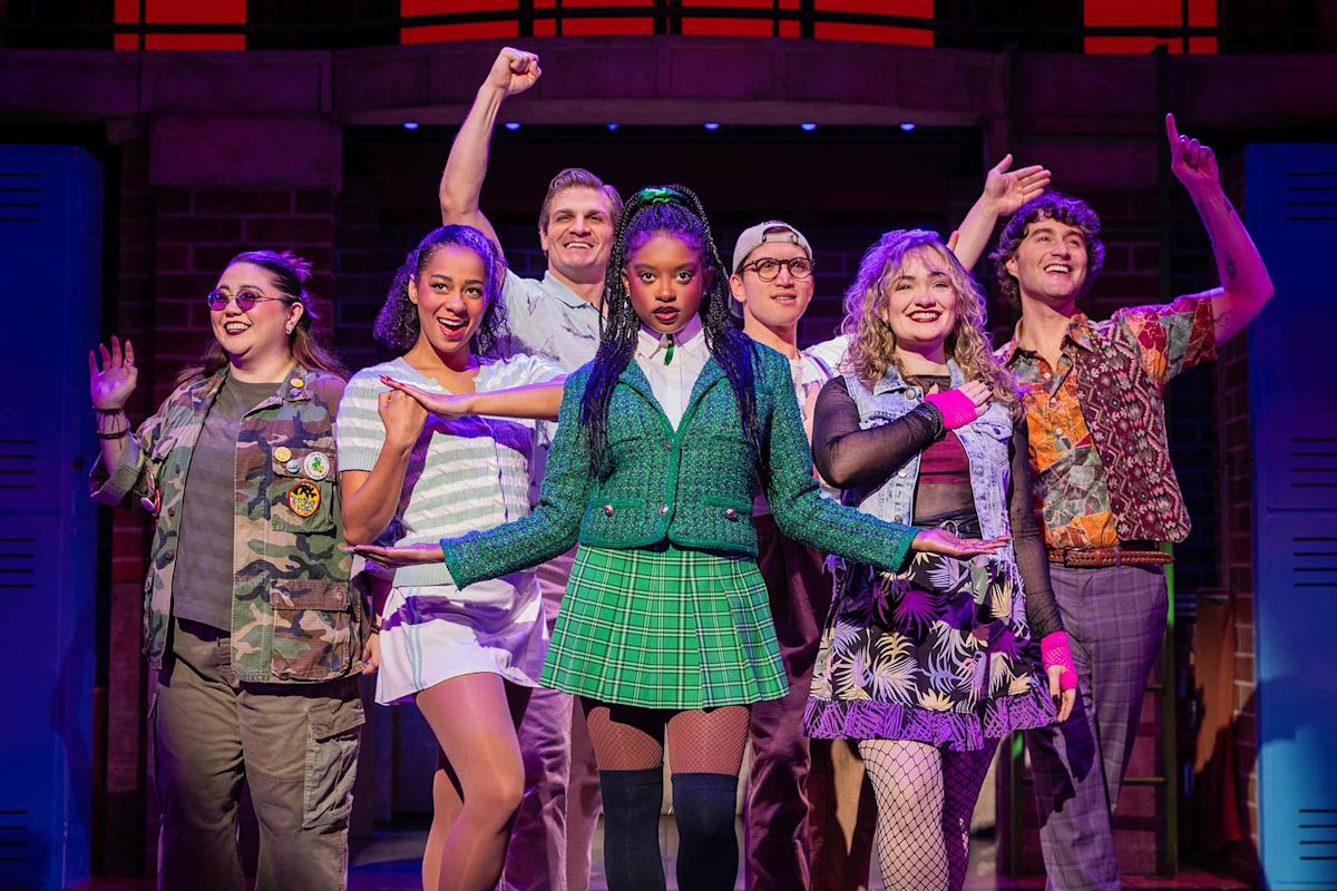 Seven actors in colorful costumes pose on stage, with the central character in a green plaid skirt and jacket, standing confidently as others surround her in expressive stances, under vibrant stage lighting.