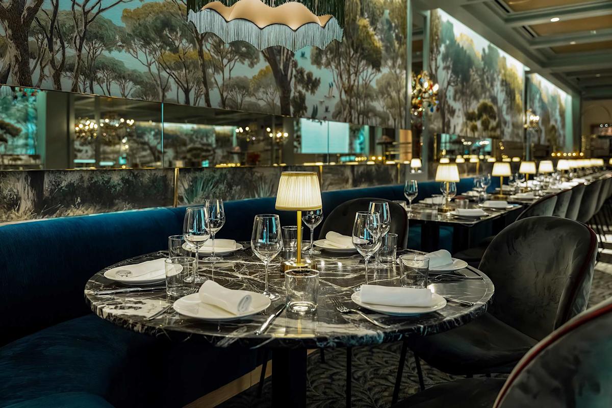 A stylish restaurant interior with marble tables set with glasses, plates, and napkins. Blue velvet seating lines the wall, and small lamps provide warm lighting. Wall mirrors reflect scenic murals of trees.