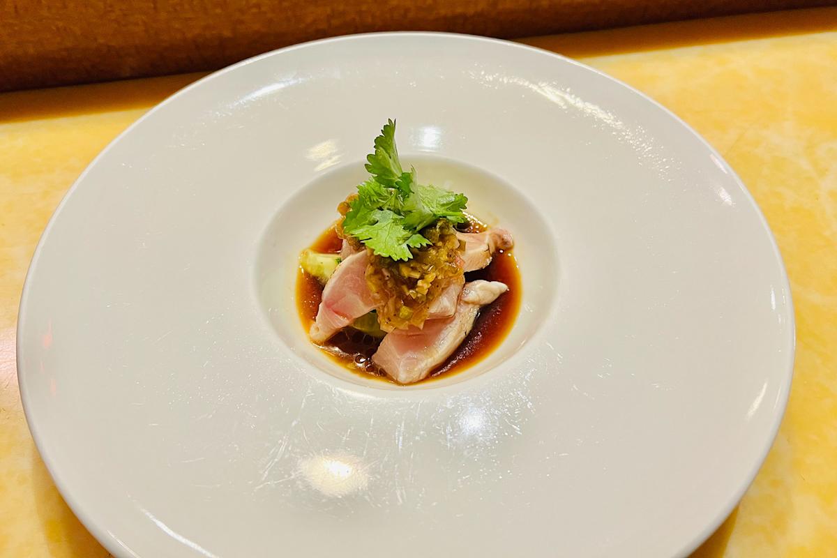 A small serving of sashimi topped with sauce and fresh cilantro sits on a bed of dark soy-based sauce, plated on a large white dish.