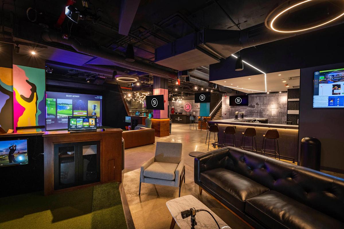 Modern indoor golf lounge with virtual golf simulators, a sleek bar with stools, colorful wall art, and comfortable seating including a black leather sofa and armchairs, all under stylish, circular ceiling lights.