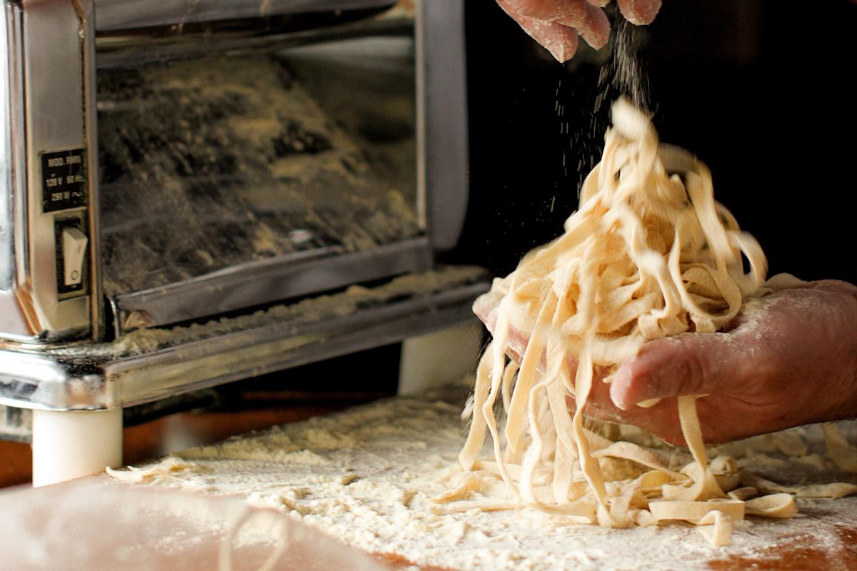 A close-up of hands holding freshly cut pasta noodles on a floured surface, with a metal pasta machine in the background and flour being sprinkled over the noodles.