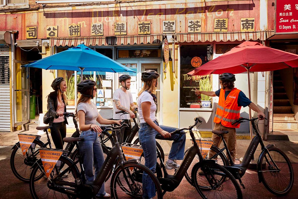 Five people wearing helmets stand with bicycles under colorful umbrellas on a street in front of a building with Chinese signage. One person in a reflective vest appears to be guiding or instructing the group.