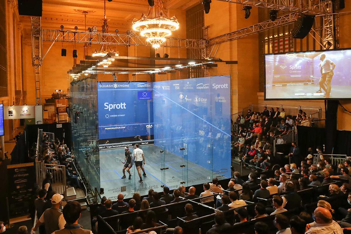 A squash match takes place in a glass court surrounded by a seated audience in a large, elegant hall with chandeliers. A large screen displays the match above the crowd.