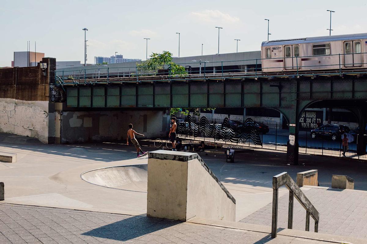 Two people are skateboarding under a green bridge at an urban skatepark as a subway train passes overhead. Concrete ramps and rails are visible, with city buildings in the background.