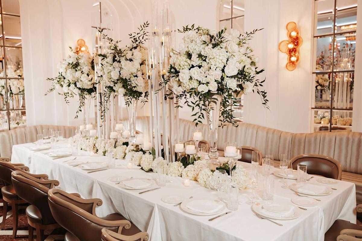 Elegant dining table set for a formal event, decorated with tall glass vases of white flowers and greenery, surrounded by beige chairs, white linens, and softly lit candles in a luxurious room.
