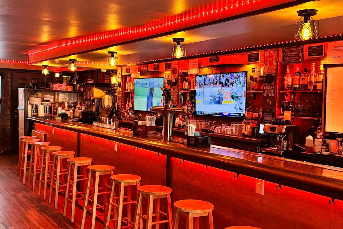 A warmly lit bar with red lighting, wooden stools lined up at the counter, two large TVs showing sports, and shelves stocked with bottles and glasses in the background.