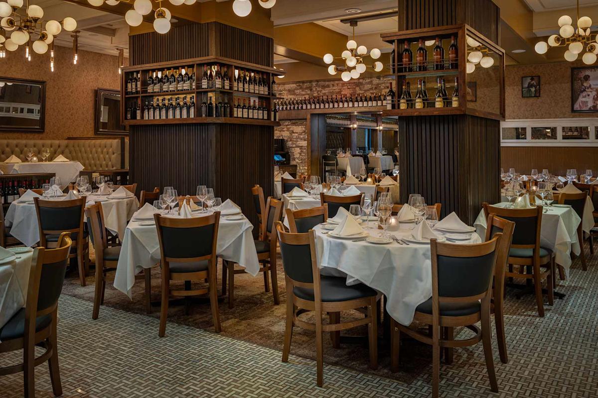 Elegant restaurant dining room with neatly arranged tables covered in white cloths, set with glasses and cutlery. Wooden chairs surround the tables, and wine bottles are displayed on shelves between pillars. Warm, ambient lighting.