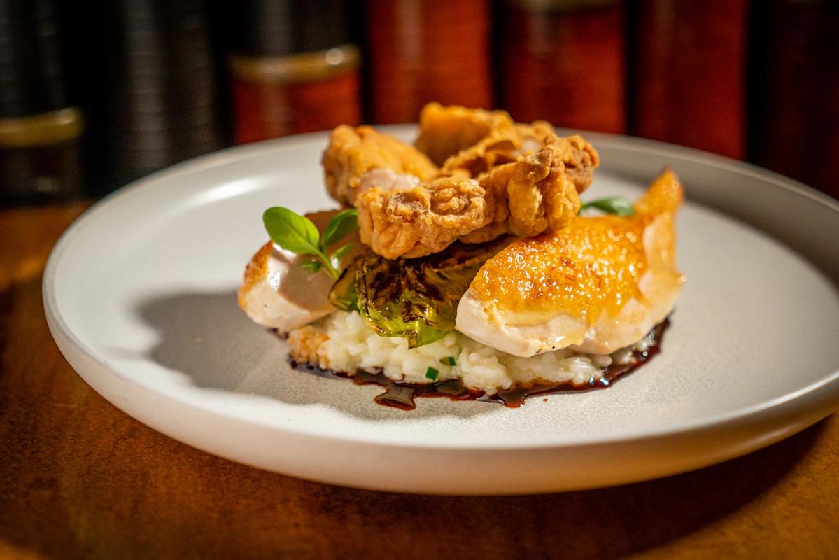 A gourmet dish featuring crispy fried chicken atop roasted chicken breast, served on creamy risotto with greens, garnished with sauce on a white plate.