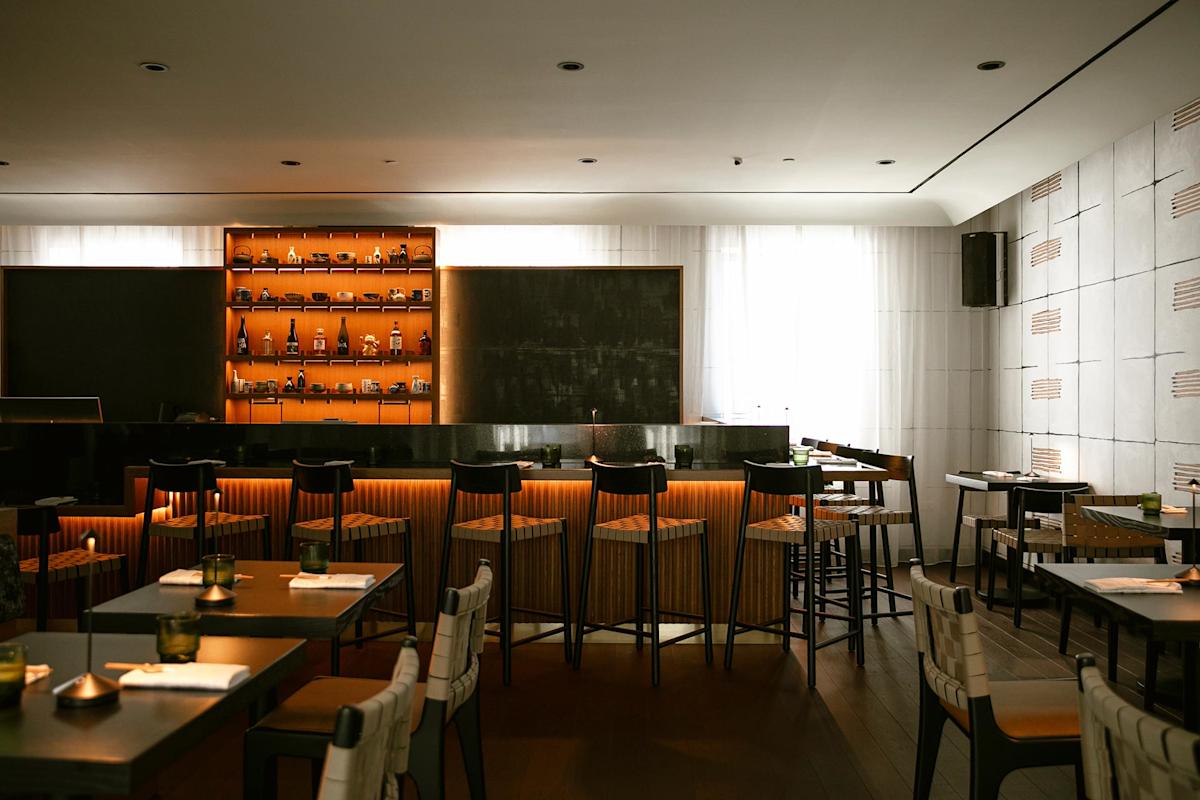 A modern, stylish restaurant interior with a bar area featuring shelves of bottles, high chairs, and warm lighting. Tables and chairs are neatly arranged, and natural light filters through sheer curtains.