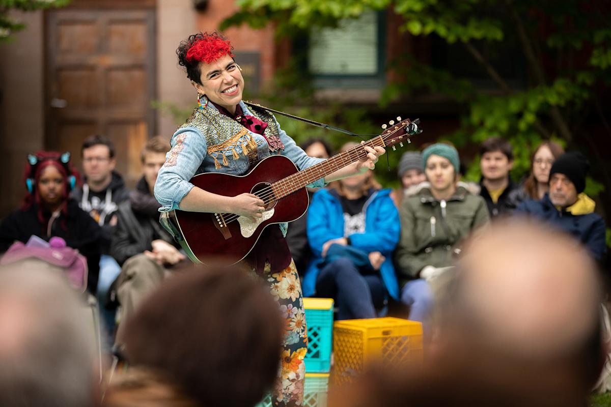 A musician with short curly red and black hair plays an acoustic guitar and smiles at an outdoor event, with an audience seated behind them. Trees and a building are visible in the background.