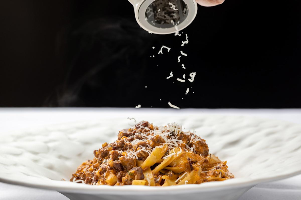 A close-up of pasta with meat sauce in a white bowl, as grated cheese is being sprinkled from above, set against a dark background.