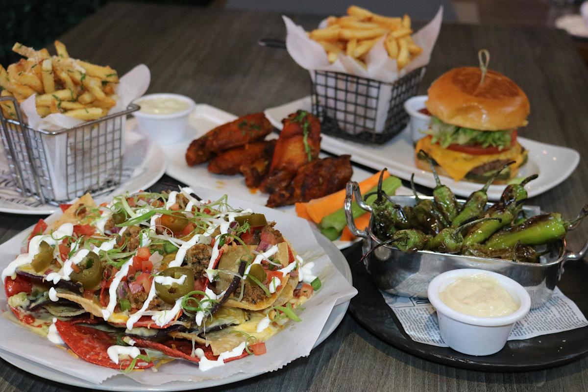A table filled with loaded nachos topped with jalapeños and sour cream, shishito peppers with dipping sauce, a cheeseburger with lettuce and tomato, two baskets of fries, and a plate of glazed chicken wings.