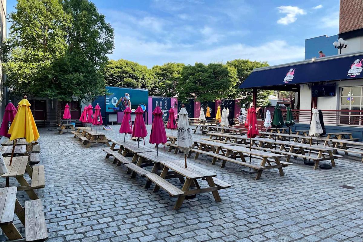 Outdoor seating area with long wooden picnic tables and closed colorful umbrellas. The ground is paved with cobblestones, surrounded by trees and buildings under a blue sky with scattered clouds.