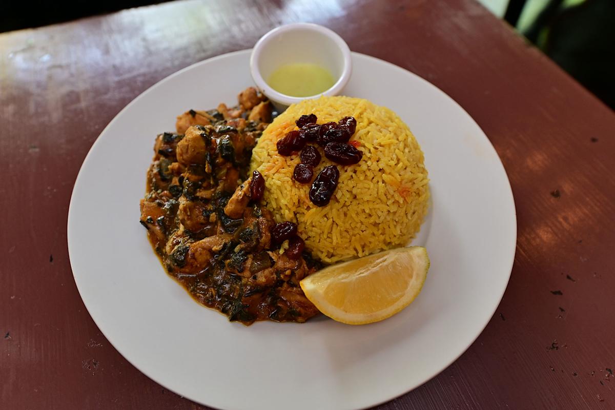 A white plate with a mound of yellow rice topped with dried cranberries, a serving of chicken and spinach curry, a lemon wedge, and a small cup of green sauce on the side.