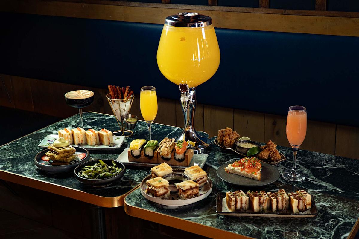 A marble table filled with assorted Japanese small plates, sandwiches, fried chicken, edamame, cocktails, and a large glass dispenser of orange drink in the center. The setting is modern and stylish.