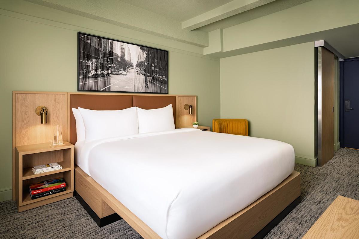 A modern hotel room with a neatly made double bed, wooden headboard and nightstands, a glass of water, books, an orange chair, and a black-and-white city street photo above the bed.