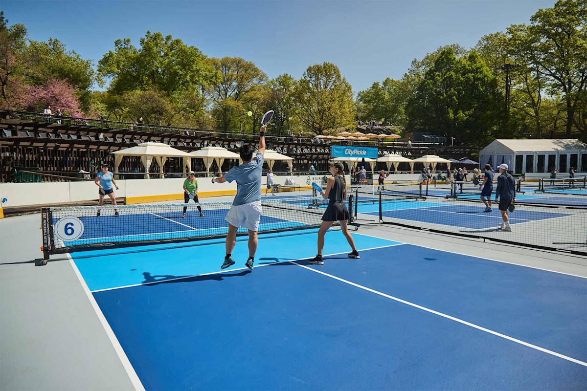 People play pickleball on outdoor blue courts under a clear sky, surrounded by trees. Two players in the foreground, one in mid-air, compete at the net while others play on nearby courts. Spectators watch from shaded seating.