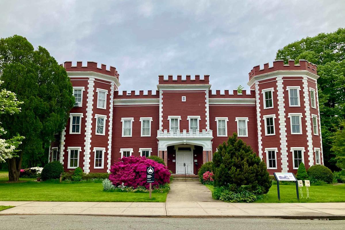 A large, red building with white trim and castle-like towers stands behind a sidewalk, surrounded by green trees, grass, and blooming pink bushes.