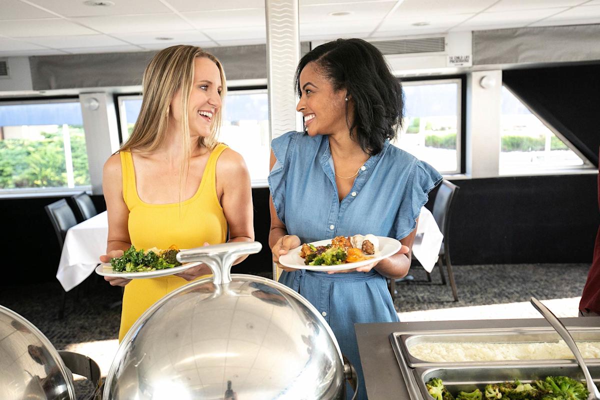 Two women smile at each other while holding plates of food at a buffet in a bright, modern dining area with large windows and white tablecloths.