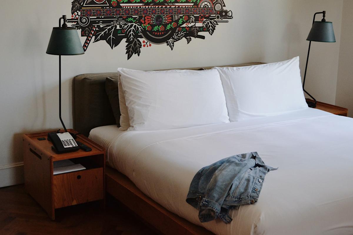 A neatly made bed with white linens and two pillows, flanked by wooden nightstands with lamps. A phone sits on the left nightstand, and a denim jacket is draped across the end of the bed.