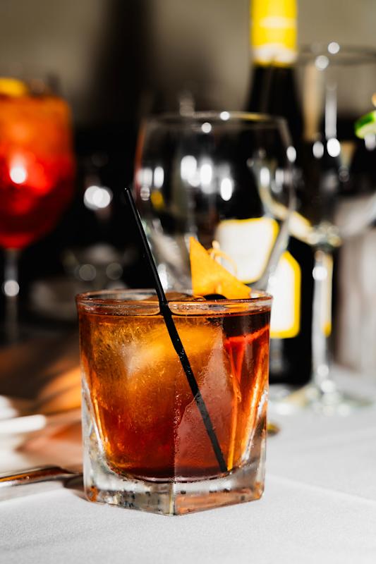 A cocktail with ice and an orange garnish in a short glass with a black straw, set on a white tablecloth with wine glasses and a bottle in the blurred background.