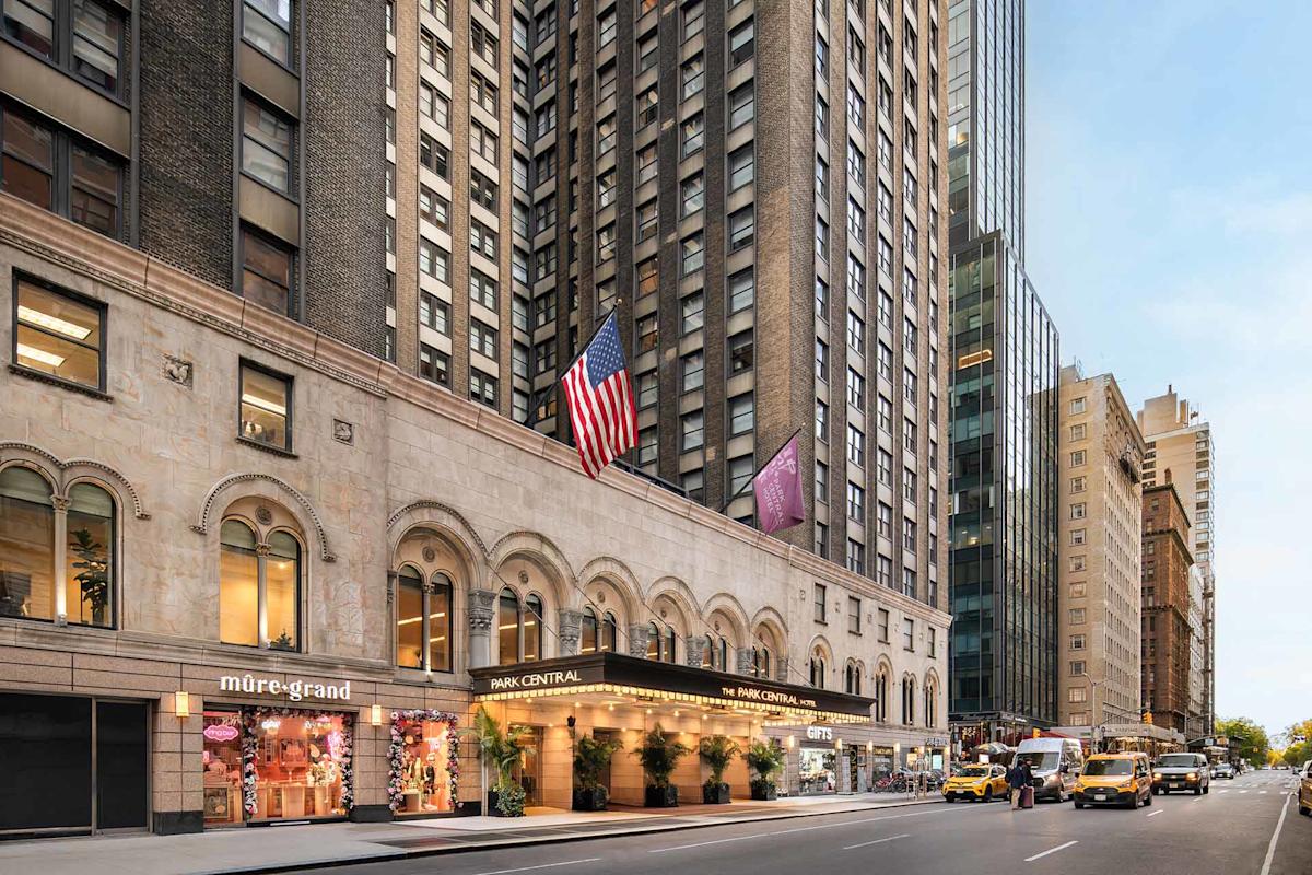 Street view of a tall hotel building with American flags, shops including "mure+grand," and yellow taxis parked along the curb in a busy city, likely New York.