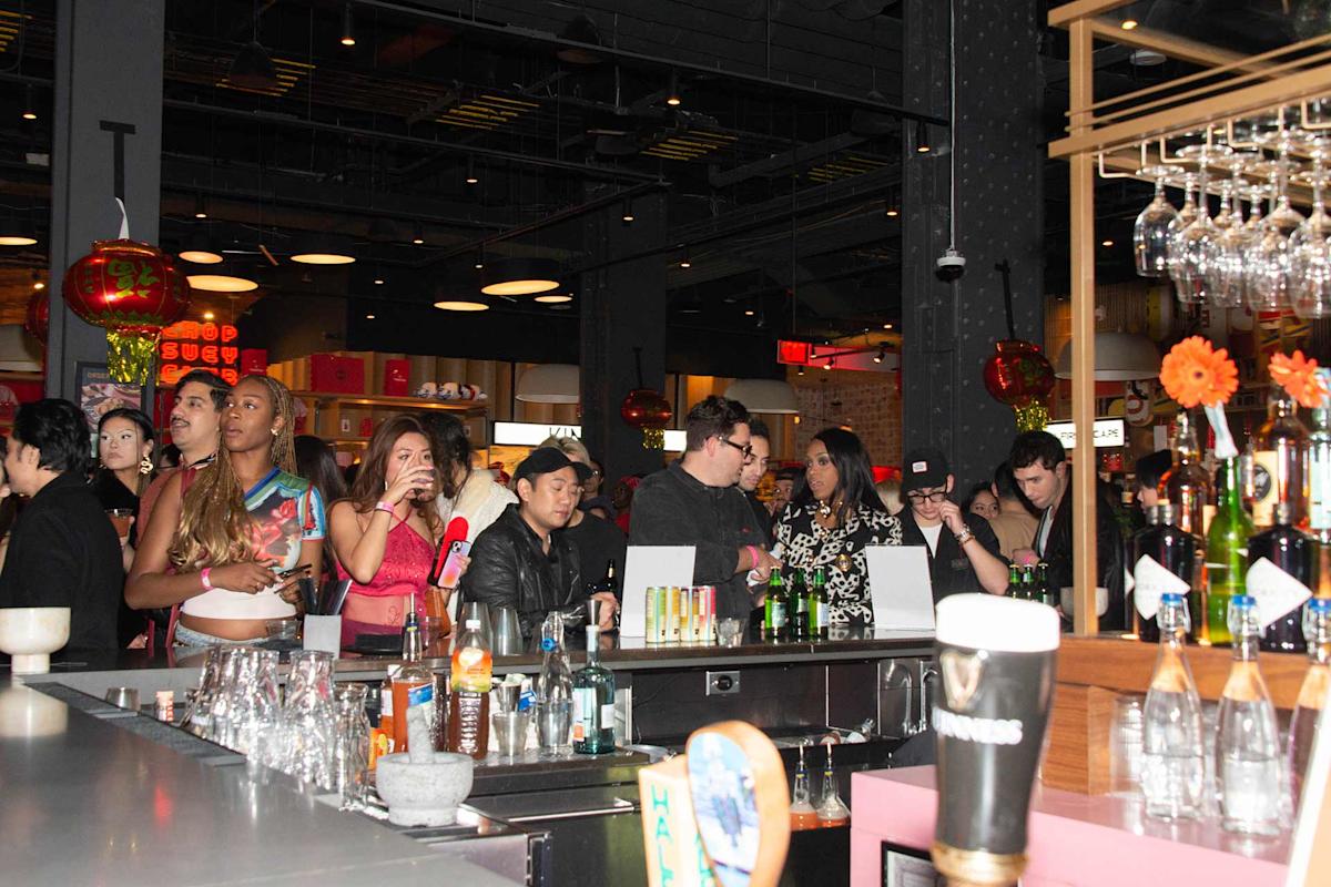 A group of people stand and socialize at a lively bar, with drinks and glassware on the counter. Red lanterns and ambient lighting decorate the background, creating a festive atmosphere.