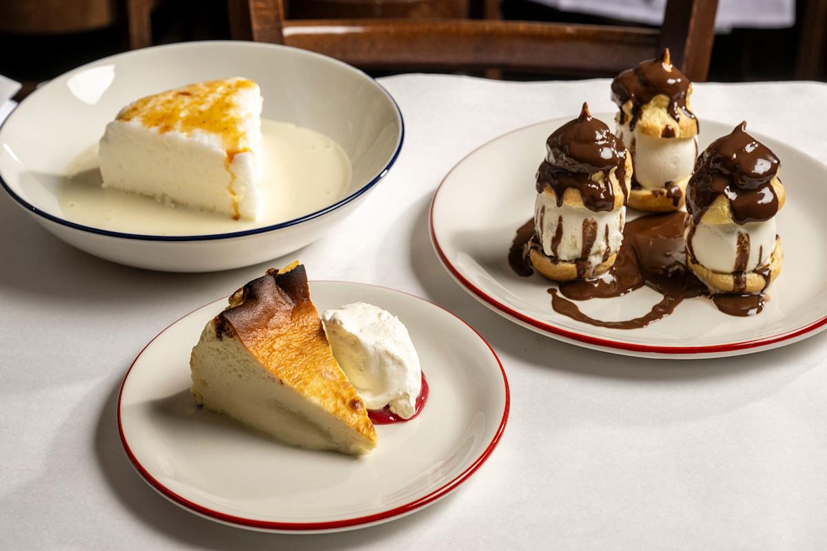 Three plates of desserts on a table: a slice of cake with sauce, three cream puffs topped with chocolate sauce, and a slice of cheesecake with a dollop of whipped cream.