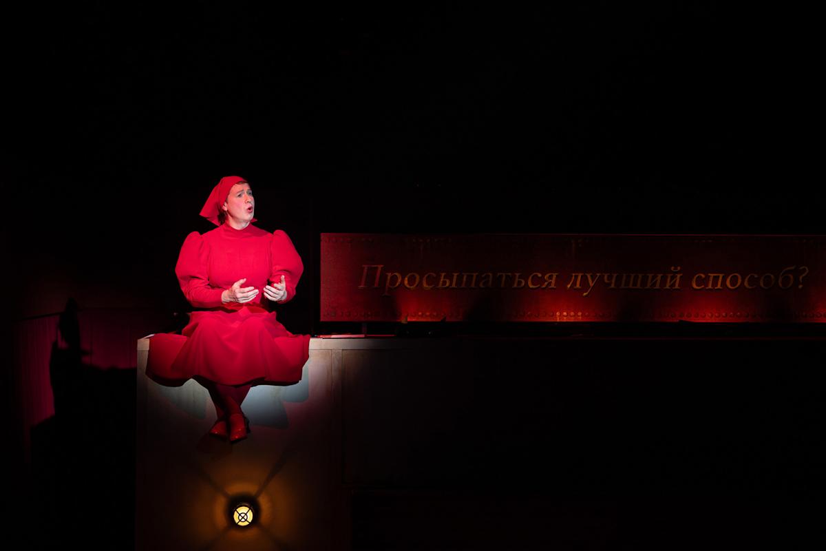 A person in a bright red costume and hat sits on a ledge, illuminated by spotlight against a dark background. Behind them, Russian text reads, "Просыпаться лучший способ?" ("Is waking up the best way?").
