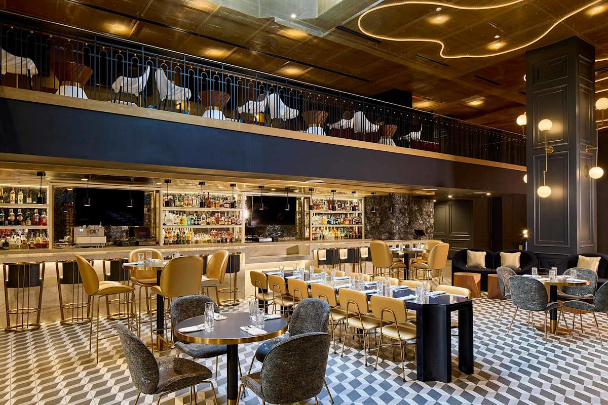 Modern, upscale restaurant interior with geometric tile flooring, gold and gray furnishings, a long bar with bottles on display, and an upper mezzanine level with additional seating. Elegant lighting fixtures hang from a gold ceiling.