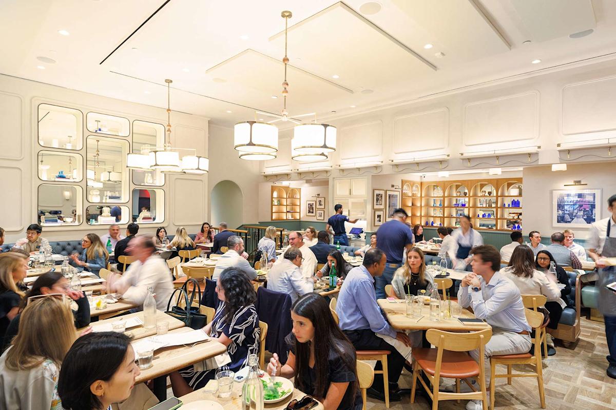 A busy, modern restaurant filled with people dining at tables. Warm lighting, white walls, and decorative shelving create a lively, inviting atmosphere. Servers move between tables as guests enjoy food and conversation.