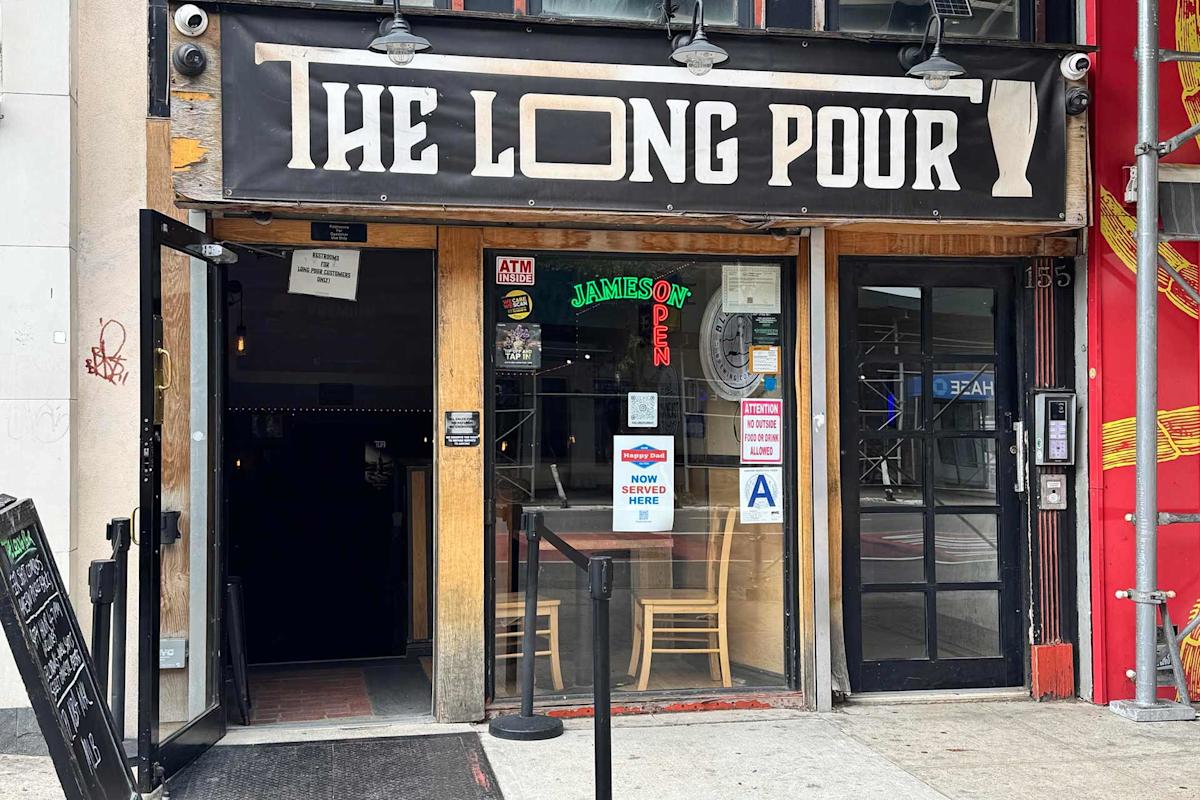 Storefront of “The Long Pour” bar with open double doors, a “Jameson” neon sign, and posters on the glass, including a “Now Served Here” beer sign. Sidewalk seating and a menu board are visible outside.