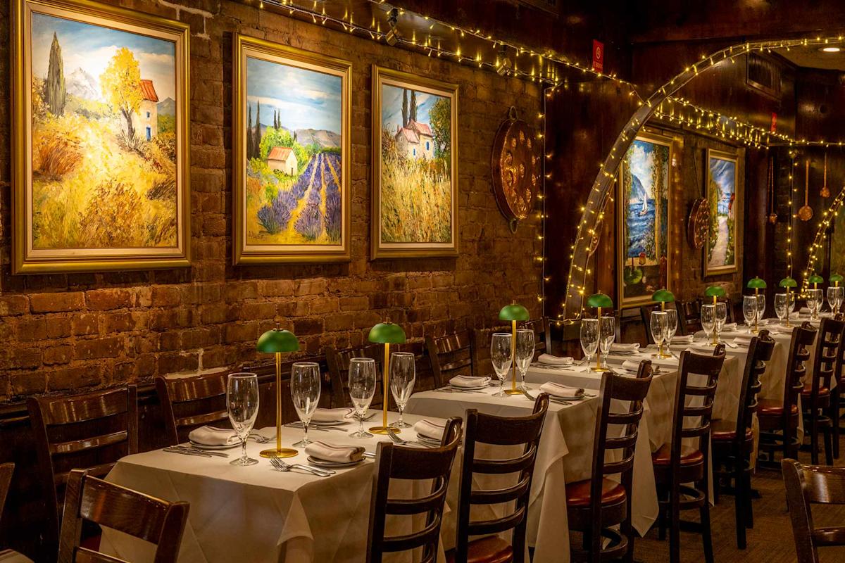 A warmly lit restaurant with white tablecloths, wooden chairs, wine glasses, and green-shaded lamps. Colorful landscape paintings hang on a brick wall adorned with string lights.
