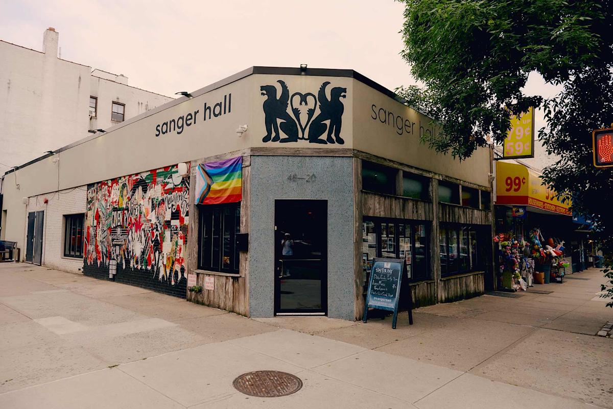 Corner view of Sanger Hall, a bar with black lion silhouettes and a heart sign on the exterior, a wall covered in posters and a rainbow flag, and a chalkboard sign by the entrance; a 99-cent store is nearby.