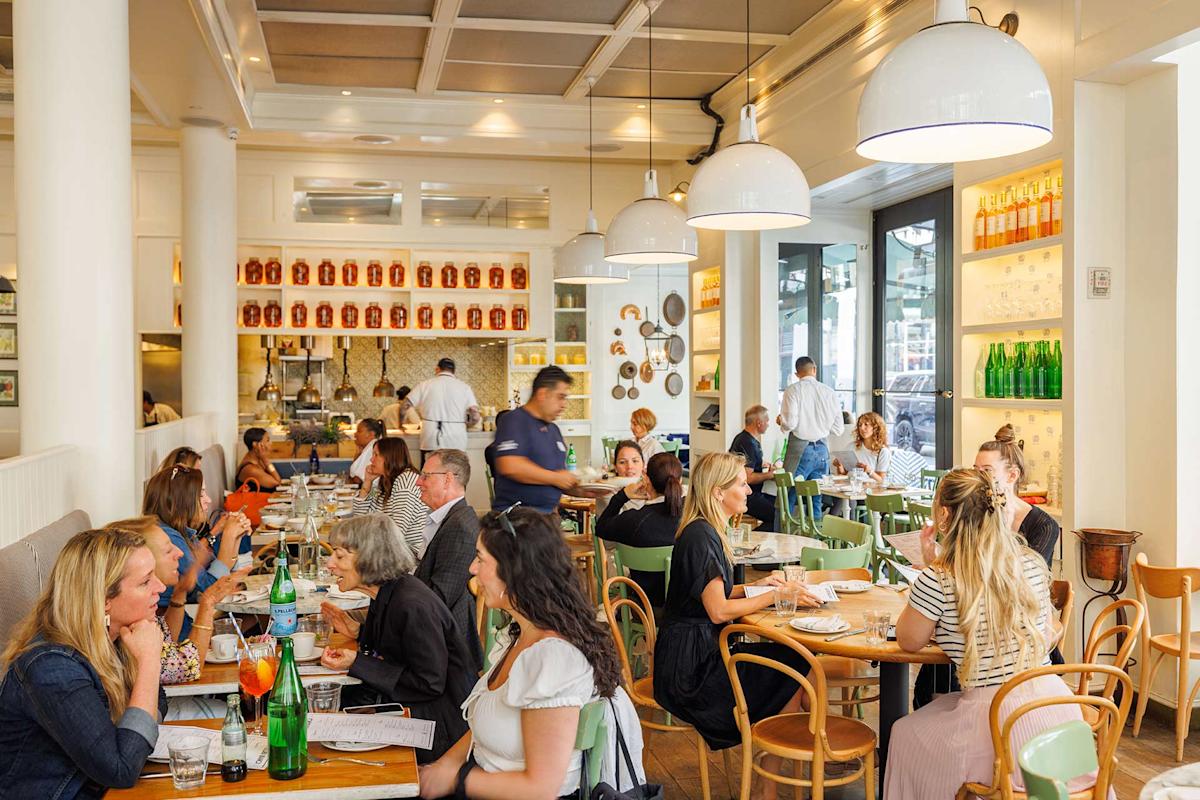 A bright, busy restaurant with people dining at wooden tables, talking and eating. Large windows let in natural light. Decorative bottles and plates line the shelves. Servers move between tables, attending to guests.