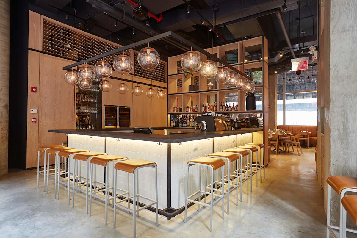 Modern bar with light wood paneling, globe pendant lights, white tiled bar counter, and tan leather stools. Shelves hold bottles and glassware; tables are visible in the background. Large windows let in natural light.