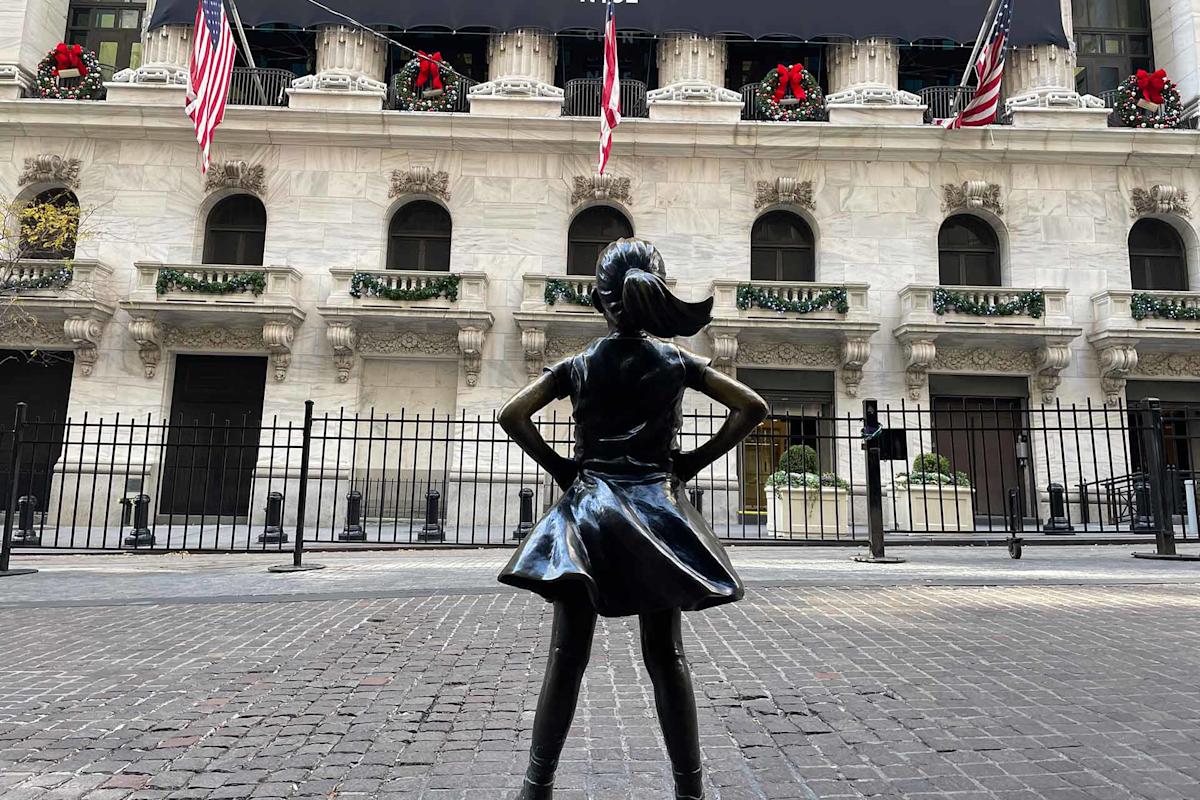 Bronze “Fearless Girl” statue stands with hands on hips facing the New York Stock Exchange, decorated with festive wreaths, American flags, and garlands on the building’s facade.