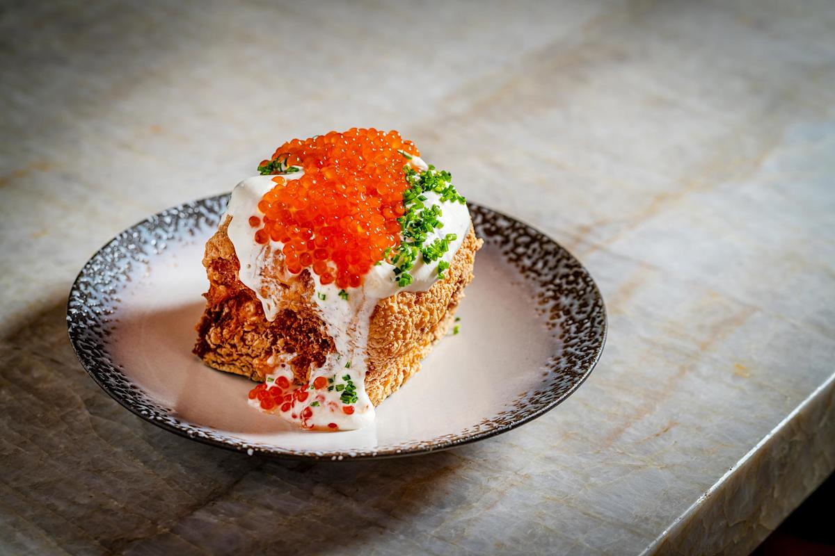 A slice of crispy bread topped with creamy white sauce, bright orange fish roe, and chopped green herbs, served on a speckled plate on a marble surface.