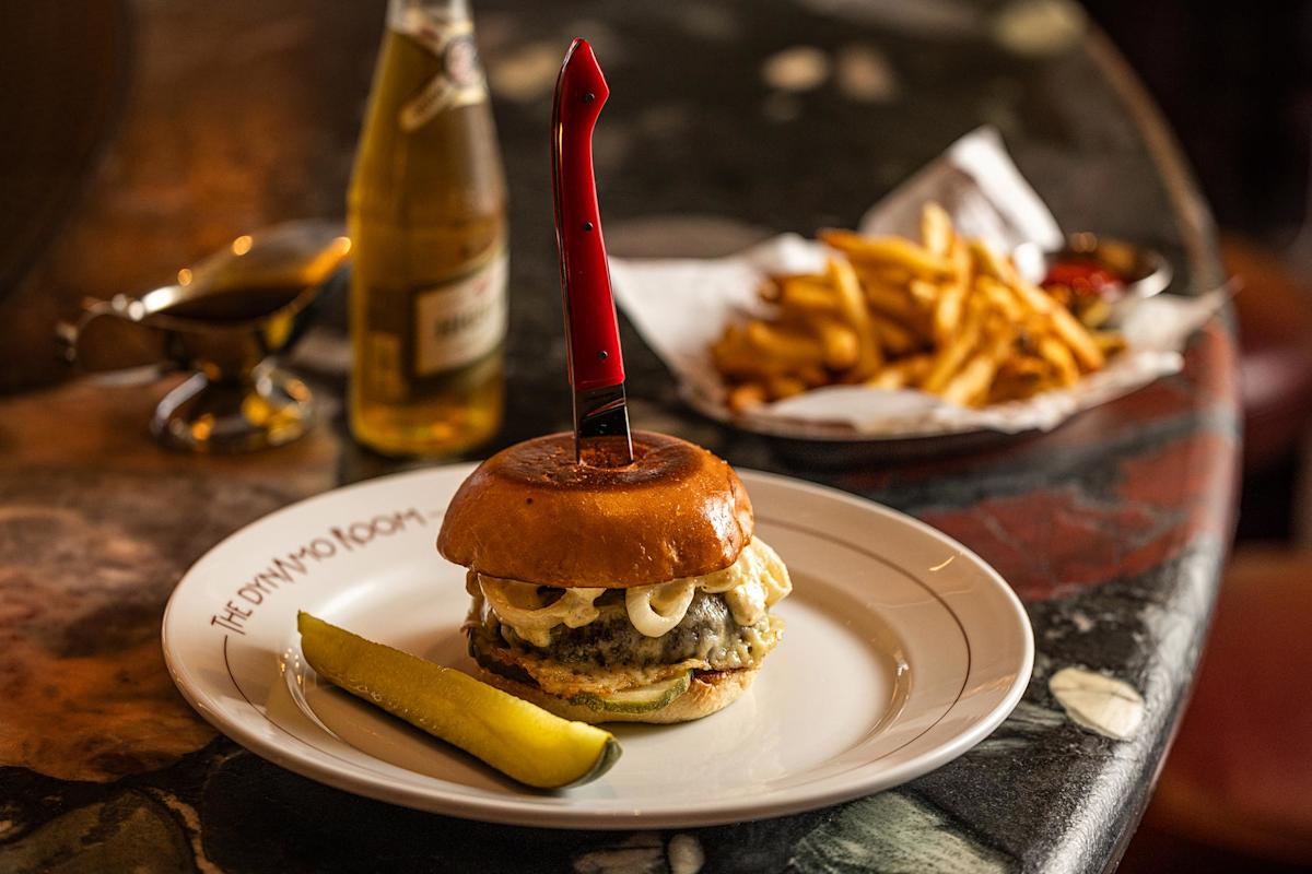 A cheeseburger with a red-handled knife through the bun is served on a plate with a pickle. In the background, there are French fries in a basket, a bottle of beer, and sauce on a marble table.