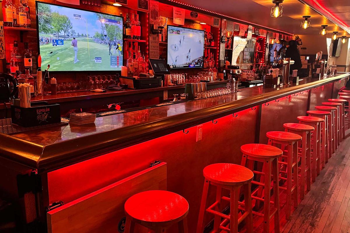 A long bar with red lighting, wooden stools lined up, and two large TVs showing sports above the counter. Glasses, bottles, and bar equipment are visible on the shelves behind the bar.