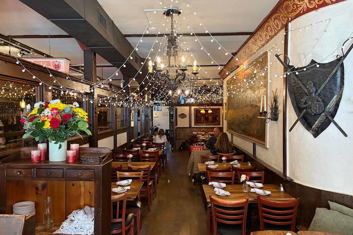 A cozy restaurant interior with wooden tables and chairs, string lights hanging from the ceiling, a shield and swords on the wall, and a vase of colorful flowers on a sideboard. Several people are seated at tables.