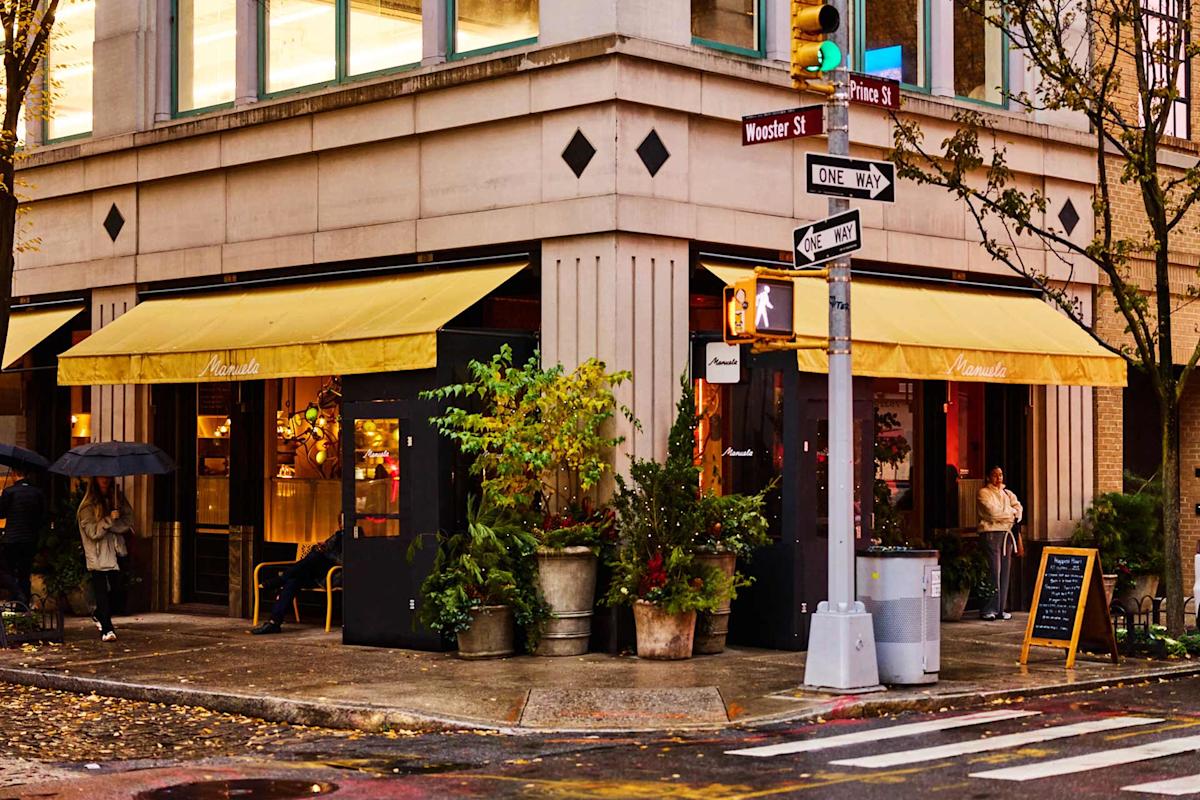 A corner café with yellow awnings and large windows, surrounded by potted plants. People stand near the entrance, and a street sign marks the intersection of Wooster St and Prince St on a rainy day.