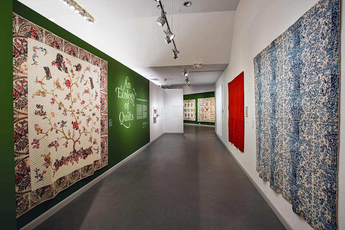 A museum gallery with colorful quilts displayed on the walls. A green wall with the text "An Ecology of Quilts" is on the left, and spotlights illuminate the quilts along the hallway.