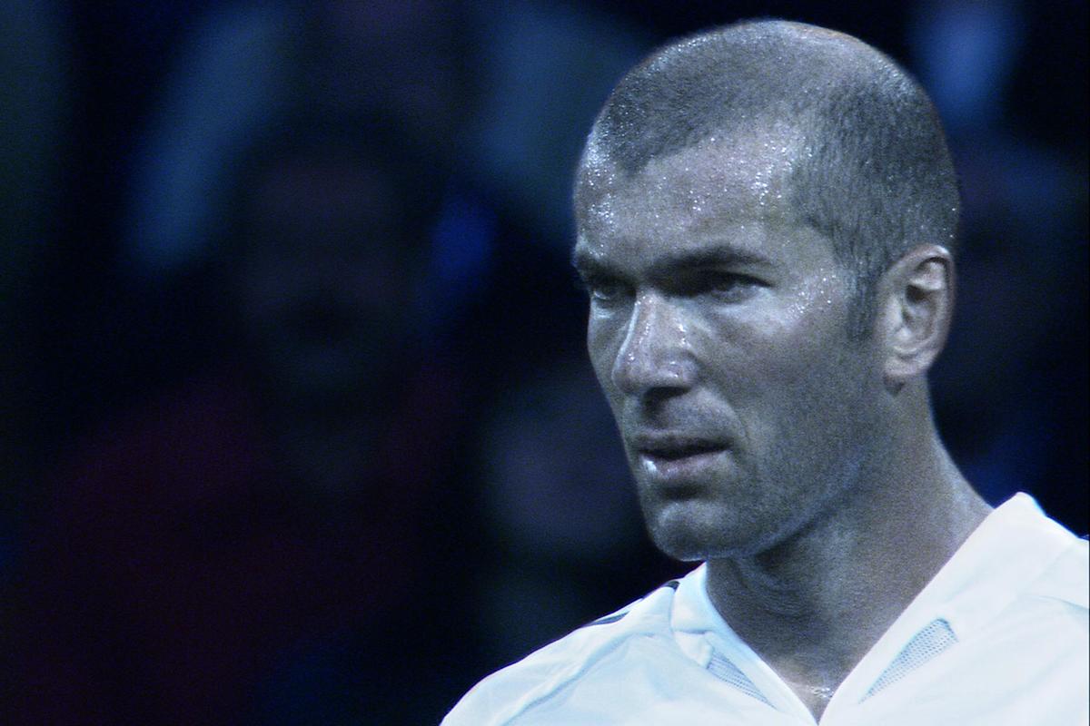 A close-up of a male soccer player with a shaved head, sweating and wearing a white jersey, looking focused. The background is blurred and dark, emphasizing his intense expression.