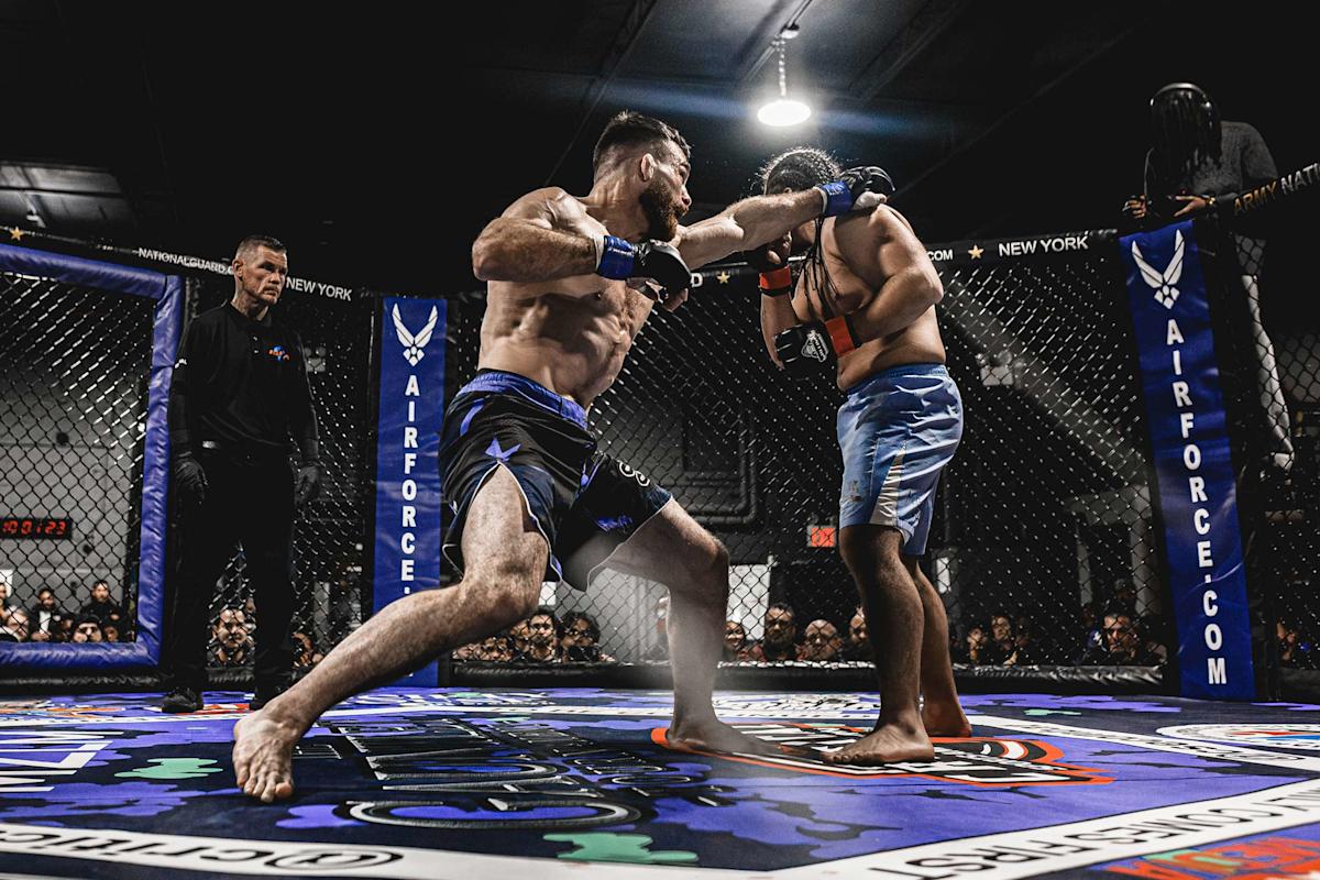 Two male MMA fighters in a cage match; one punches the other while a referee watches closely. The fighters wear blue shorts and gloves. Spectators and Air Force branding are visible around the cage.