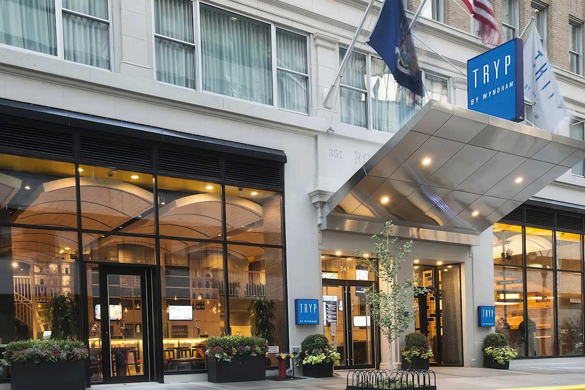 Street view of the entrance to TRYP by Wyndham hotel, featuring large windows showing the modern lobby, a silver canopy, blue hotel signs, and flags above the door.