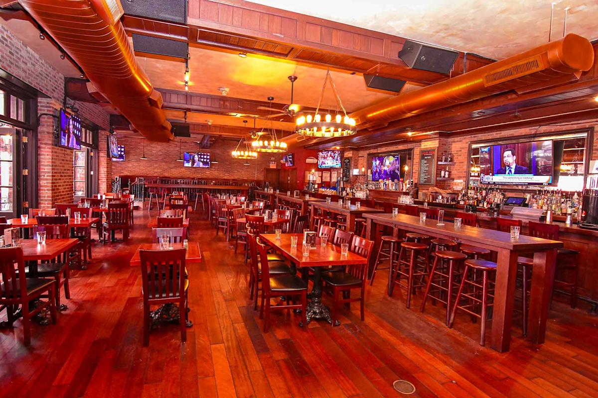 Spacious bar and restaurant with wooden floors, tables, and chairs, a long bar counter with barstools, brick walls, chandeliers, and multiple TVs showing sports and news. Warm lighting gives a cozy atmosphere.