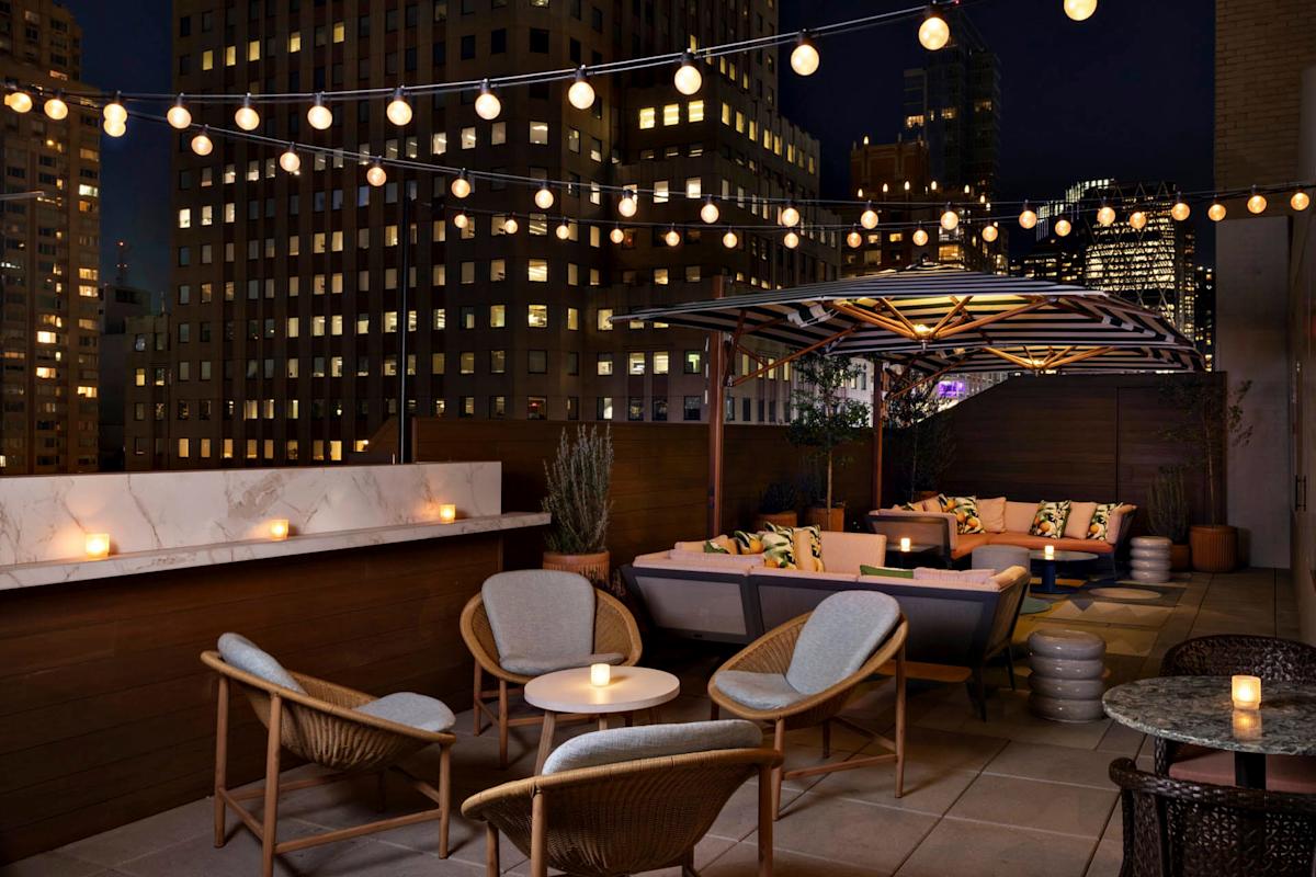 A stylish rooftop lounge at night with cozy seating, round tables, a marble-topped bar, string lights overhead, and city skyscrapers in the background. Candles on tables create a warm, inviting atmosphere.